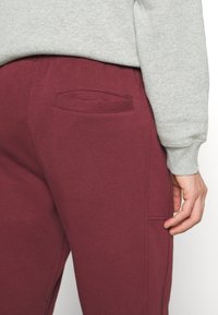 Bordeauxfarbene Jogginghose aus weichem Stoff, mit einer aufgesteppten Kontur an der Gesäßtasche und Details an der Seitennaht. Kombiniert mit einem grauen Sweatshirt.