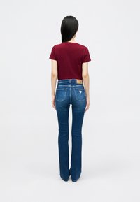 Žena s dugom crnom kosom stoji okrenuta leđa, obučena u kratkosvjetlu burgundsku majicu i plave traperice s visokim strukom i širokim nogavicama.