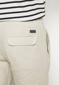 Beige linnebyxor med slim fit, som har två bakfickor och ett litet varumärkesmärke. Bärs över en svart och vit randig skjorta.