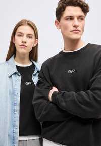 Νεαρή γυναίκα φοράει μαύρο πουκάμισο και ανοιχτόχρωμο τζιν μπουφάν· νεαρός άνδρας φοράει μαύρο φούτερ, και οι δύο με μικρό λευκό λογότυπο στο στήθος, ουδέτερο φόντο.