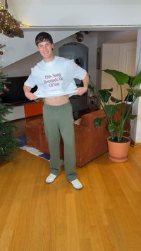 Valkoinen T-paita, jossa on burgundinpunaisella tekstillä "This Song Reminds Me Of You", yhdistetty vihreisiin collegehousuihin ja valkoisiin lenkkareihin, seisomassa sisätiloissa.