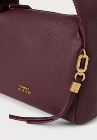 Mørk lilla teksturert lærhåndveske med gullfarget beslag og "Tommy Hilfiger"-logo preget på forsiden.