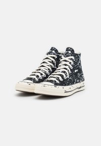 Sapatilhas de cano alto com um design salpicado em preto e branco, material de lona, atacadores em creme e sola de borracha; detalhe contrastante na biqueira.