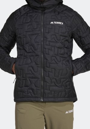 Czarna pikowana kurtka z przednim zamkiem błyskawicznym i logo Terrex, noszona na khaki spodniach również z oznaczeniem Terrex, ręce w kieszeniach kurtki.