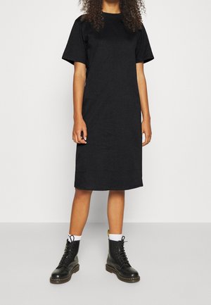 Personne portant une robe t-shirt noire longueur genoux avec manches courtes, chaussettes blanches et bottines noires à lacets, debout devant un fond uni.