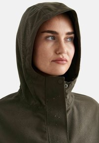 Femme portant une veste de pluie à capuche vert foncé avec des gouttes d'eau visibles à la surface, regardant sur le côté sur un fond uni.