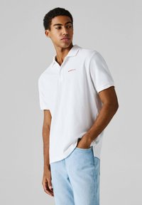 Νέος άνδρας που φοράει λευκό πόλο μπλουζάκι και ανοιχτόχρωμο τζιν, κοιτάζοντας προς τα πλάγια με το ένα χέρι στην τσέπη, μπροστά σε απλό γκρι φόντο.