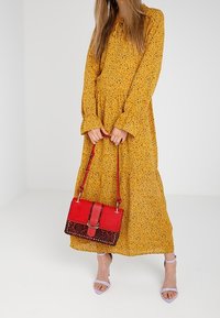 Vestido floral amarillo con mangas largas y dobladillo con volantes. Combinado con un bolso rojo con detalles decorativos y una correa para el hombro.