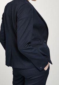 Blazer navy in tessuto testurizzato, chiusura a un bottone e due tasche laterali. Vestibilità sartoriale, spalle strutturate e dettagli di cucitura discreti.