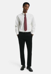 Weißes Hemd mit langen Ärmeln, kombiniert mit schwarzen Cordhosen und einer weinroten gemusterten Krawatte. Schwarze Slipper runden den Look ab.