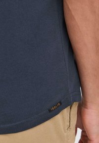 Mørkeblå bomulds t-shirt med korte ærmer, der har en lige kant og et lille sort mærke med "ISOLID" i orange på side sømmen.