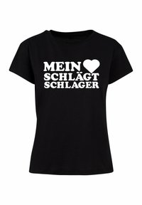 Sort T-shirt lavet af bomuld med fed hvid tekst, der står "MEIN ❤️ SCHLÄGT SCHLAGER" på forsiden. Korte ærmer og rund hals.
