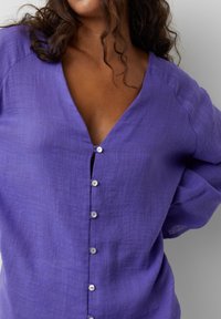 Blusa de lino morada con escote en V, mangas largas y cinco botones en el frente. Tela texturada y corte relajado.