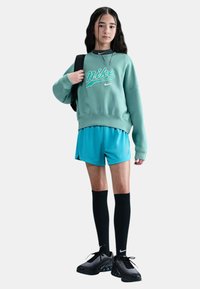 Giovane che indossa un felpa Nike verde, pantaloni corti blu, calze nere fino al ginocchio, scarpe da ginnastica nere, mentre porta uno zaino nero, in piedi su uno sfondo bianco.