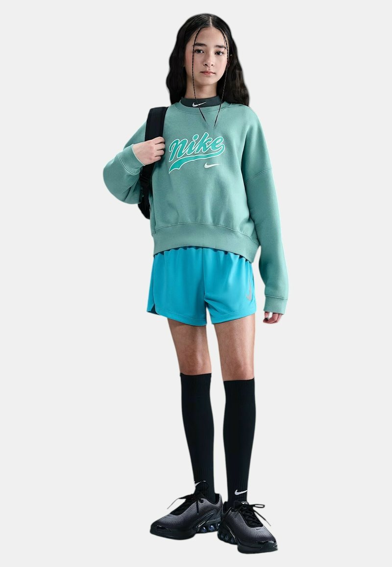 Giovane che indossa un felpa Nike verde, pantaloni corti blu, calze nere fino al ginocchio, scarpe da ginnastica nere, mentre porta uno zaino nero, in piedi su uno sfondo bianco.