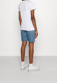 Valkoinen lyhythihainen t-paita, siniset farkkushortsit, valkoiset korkeat tennarit mustilla yksityiskohdilla, näkyvät saumat, nähtynä takaa.