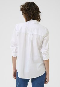 Chemise blanche à manches longues, coupe décontractée, détail de pli au dos et ourlet arrondi. Associée à un jean en denim bleu.
