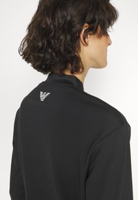 Mustassa polo-paidassa on sileä rakenne, ja sen takana on pieni valkoinen logo. Kaulus on litteä ja muotoilu on minimalistinen.