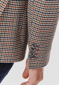 Wzorzysty rękaw marynarki w deseń houndstooth w odcieniach brązu, granatu i czerwieni, z czarnymi guzikami i subtelnym detalem kieszeni.
