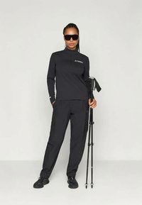 Μαύρη μπλούζα με μακριά μανίκια, κολάρο με φερμουάρ και λογότυπο. Μαύρο παντελόνι cargo με τσέπες. Μοντέλο που κρατάει ένα μπαστούνι ορειβασίας, φορώντας σκούρα γυαλιά ηλίου.