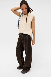 Cremefarvet strikket ærmeløs sweater med struktureret mønster, parret med løstsiddende brune kordfløjlsbukser og mørke slip-on sko.