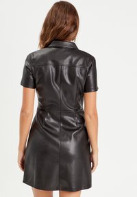 Robe chemise en cuir noir avec manches courtes, coupe ajustée et une légère brillance. Présente un col, des coutures dans le dos et une jupe évasée.