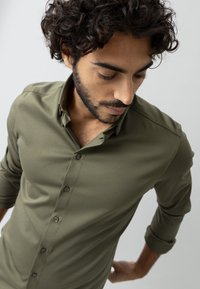 Camicia verde oliva con bottoni, maniche lunghe arrotolate, texture morbida, design aderente, leggero lucido, cinque bottoni sul davanti, colletto ordinato.