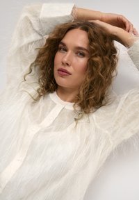 Sheer long-sleeve blouse with a textured, fuzzy appearance in off-white. Features buttons down the front, with a round neckline and soft silhouette.
