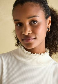 Kerge beež ribadest topp kõrge volangkraega. Mudel kannab kuldseid rõngaskõrvarõngaid. Taustal on pehme muted toon.