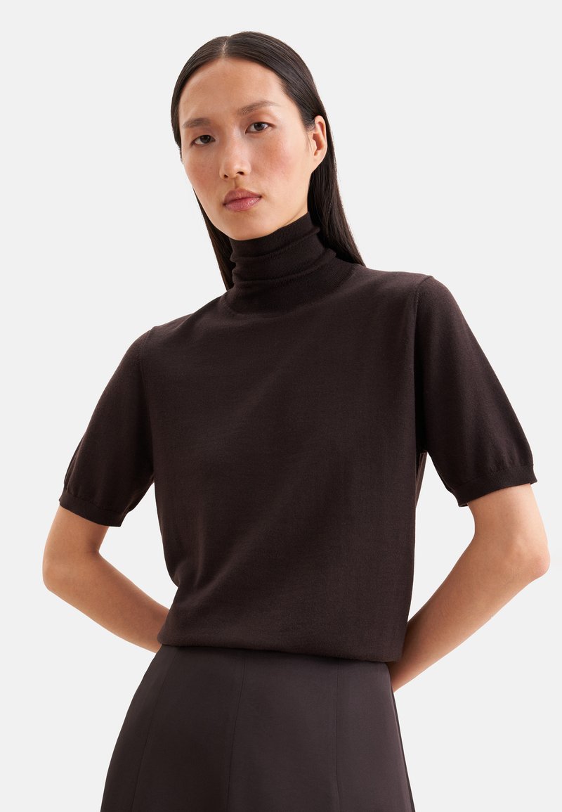 Femme aux longs cheveux foncés portant un pull col roulé noir à manches courtes et une jupe noire, debout les mains derrière le dos.