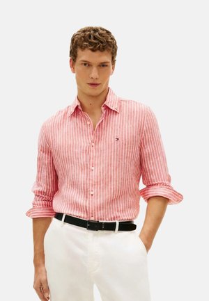 Joven con una camisa de manga larga con rayas verticales rojas y blancas, las mangas remangadas, pantalones blancos y un cinturón negro, de pie con una mano en el bolsillo.