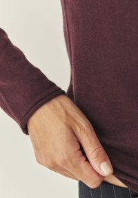 Burgundský pletený svetr s měkkou Texturou, přiléhavými dlouhými rukávy a designem manžet. Ruka uchopila lem, což ukazuje na detail a kvalitu tkaniny.
