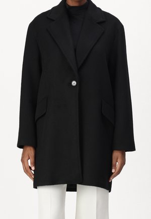 Czarny płaszcz na jeden guzik z klapami i przednimi kieszeniami z patkami, noszony na czarnej bluzce i białych spodniach, pokazany od połowy uda do szyi.