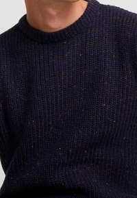 Marinblå stickad tröja med rund halsringning, ribbade ärmslut och färgglada prickar genom den strukturerade tyget.