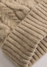 Beige gestrickte Wollmütze mit Kabelmuster und geripptem Bündchen, aus weichem, strukturiertem Stoff und in einer warmen, neutralen Farbe.