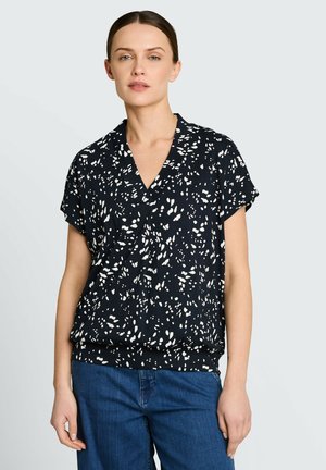 Kobieta o brązowych włosach, ubrana w czarną bluzkę z krótkim rękawem z białymi abstrakcyjnymi plamami i niebieskie dżinsy, stojąca na jasnoszarym tle.