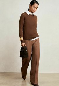 Suéter marrón de punto con dobladillo acanalado sobre una camisa blanca con cuello, combinado con pantalones de pierna ancha marrones. Sosteniendo un pequeño bolso de ante negro.