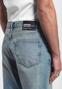 Jasnoniebieskie dżinsy mają wysoką talię, prosty krój i subtelne wypalenia. Z tyłu przymocowana jest czarna metka z białym napisem.