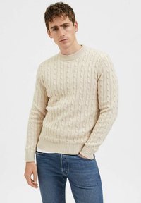 Suéter de punto beige con cuello redondo, pu�ños y dobladillo de canalé, llevado sobre una camisa blanca, combinado con jeans de mezclilla azules.