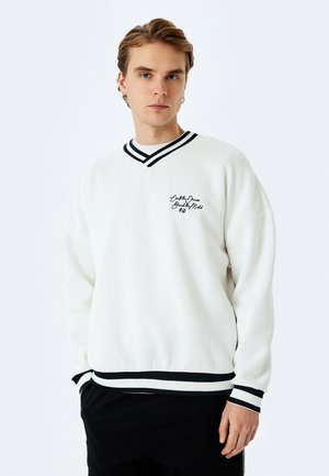 Biały sweter z czarno-białym, paskowanym dekoltem w serek, elastycznymi mankietami i dołem. Posiada teksturowany materiał oraz haftowane logo na klatce piersiowej.