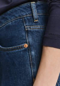 Close-up van de zak van een blauwe spijkerbroek met een koperen klinknagel en een naastliggend arm die een donkerblauwe longsleeve shirt draagt.