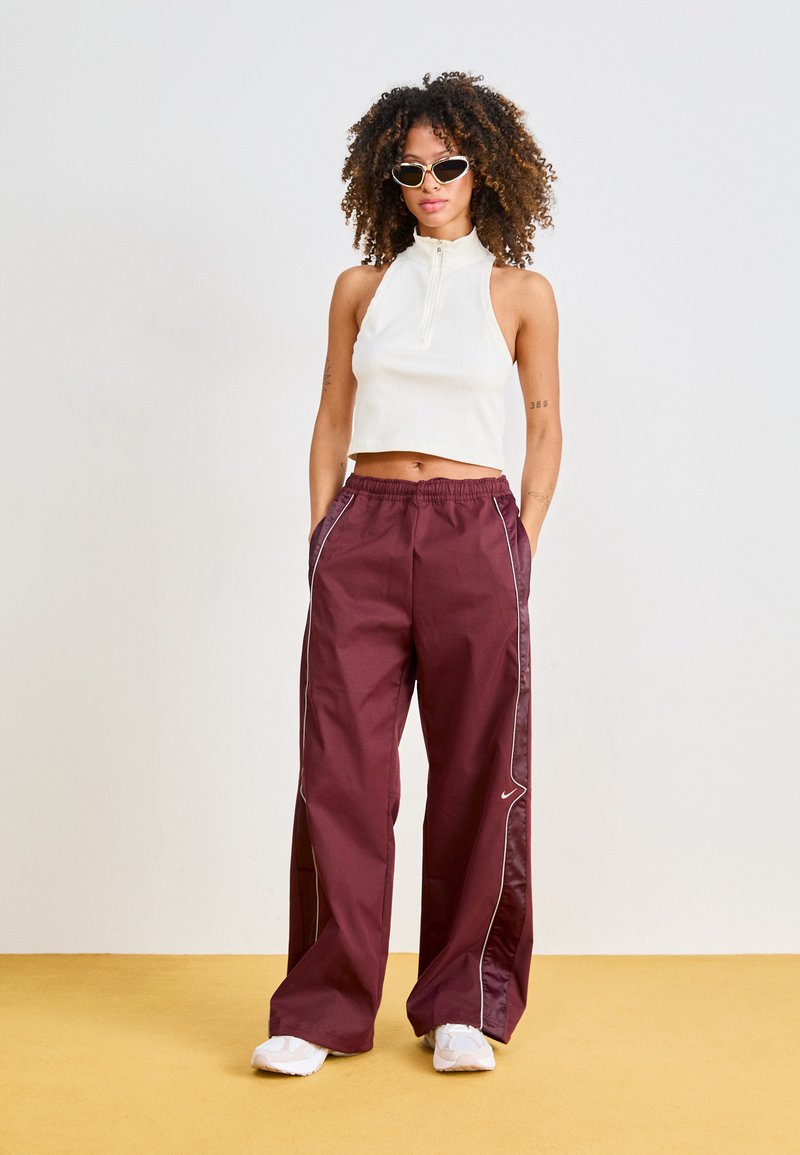 Pantalons larges bordeaux avec piping blanc, associés à un haut sans manches blanc à côtelés. Le modèle porte des baskets blanches et des lunettes de soleil.