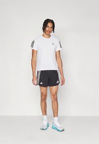 Άσπρο αθλητικό κοντομάνικο μπλουζάκι με μαύρες ρίγες, συνδυασμένο με μαύρο σορτς που φέρει λογότυπο. Φορεμένο με ανοιχτόχρωμα παπούτσια και άσπρες κάλτσες.