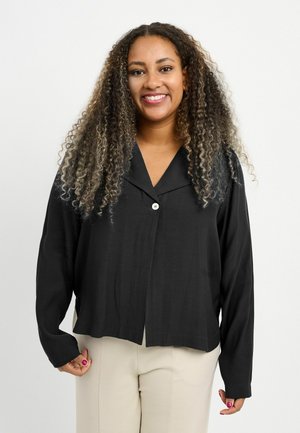 Mujer sonriente con cabello largo y rizado, que lleva una camisa negra abotonada y pantalones beige, de pie frente a un fondo blanco liso.