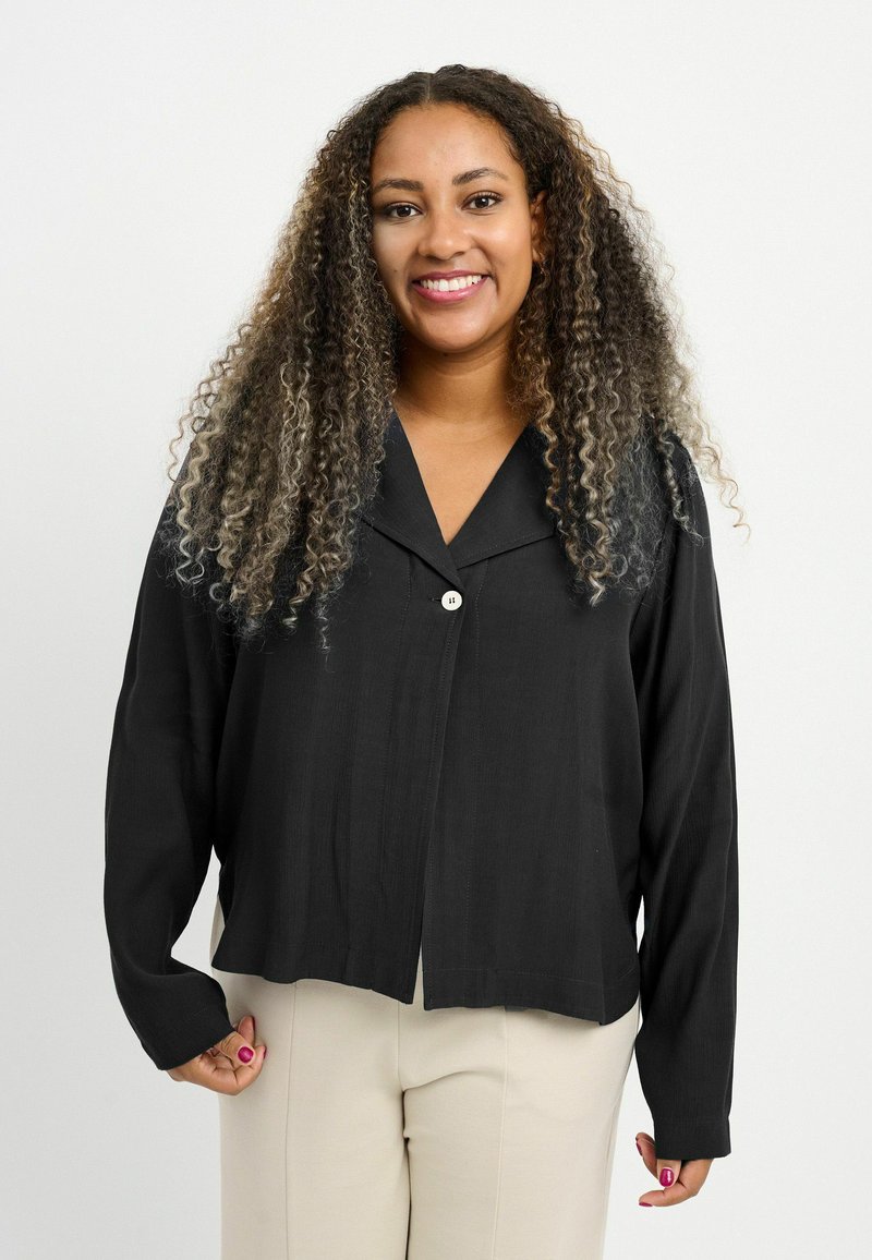 Femme souriante aux longs cheveux bouclés, vêtue d'une chemise noire boutonnée et d'un pantalon beige, debout devant un fond blanc uni.