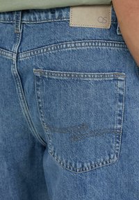 Jasnoniebieskie dżinsy z denimu z tylną kieszenią z haftowanym wzorem i brązową metką oznaczoną "QS" w pobliżu pasa.