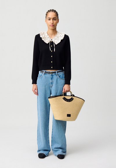Suéter negro recortado con un cuello de volantes de lunares blancos, combinado con vaqueros de pierna ancha de color azul claro y una bolsa de paja beige con detalles negros.