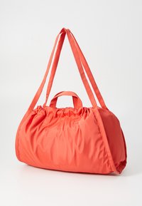 Korallfärgad tygväska med rund form, kort handtag och långa axelremmar; har en samlad toppdesign för enkel öppning och stängning.