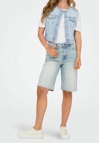 Džínová vesta se světlým praním, se zapínáním na knoflíky a dvěma náprsními kapsami, spárovaná s džínovými šortkami do kolen a bílými teniskami.