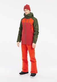 Rode en groene waterdichte jas met een rits aan de voorkant en een capuchon, gecombineerd met feloranje broeken en zwarte sneeuwlaarzen.